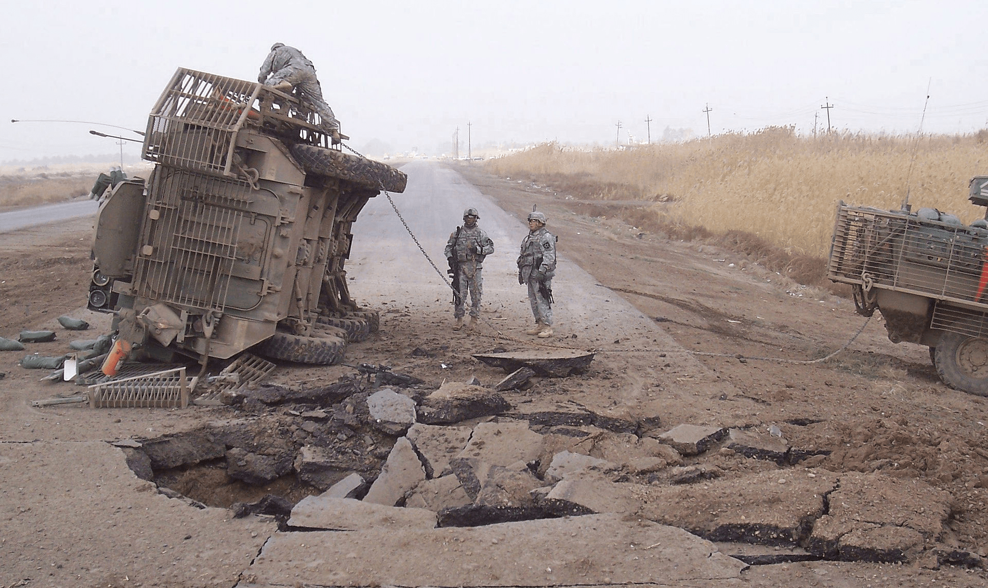 US Military standing next to an overturned tactical vehicle with a bomb blast hole next to them
