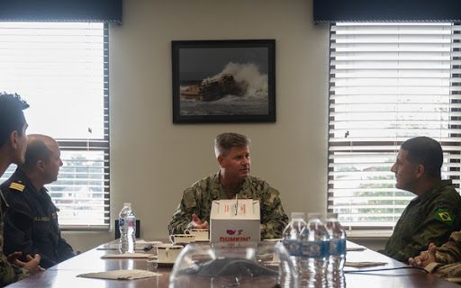 Military members sitting at a conference table holding a meeting. There is also coffee and water on the table with a painting in the background