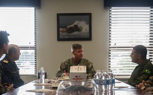 Meeting Time Military members sitting at a conference table holding a meeting. There is also coffee and water on the table with a painting in the background