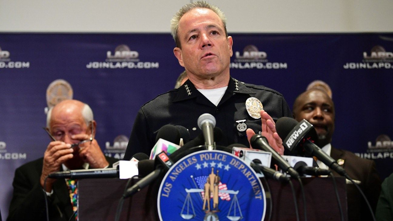 Police Chief in uniform gives press conference in front of a lectern with multiple microphones mounted on it. There is a seal of the Los Angeles Police Department on the lectern