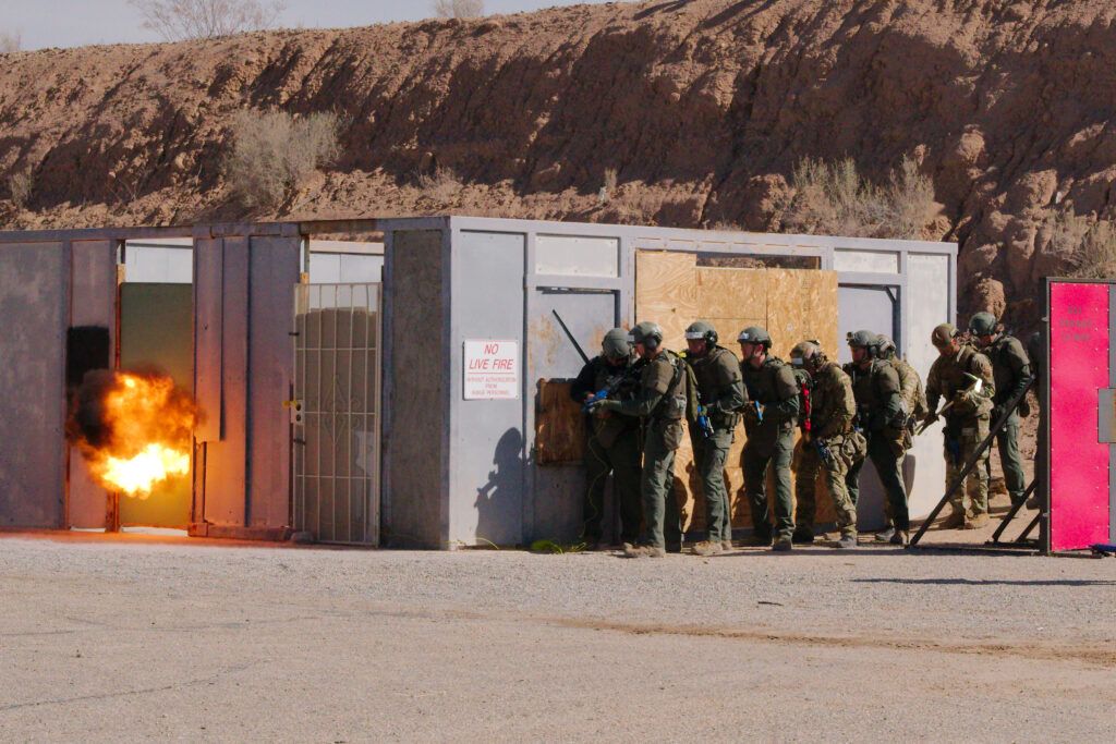 A tactical team waits around the corner of a building for an explosive breach to open the door. This is set in a training environment